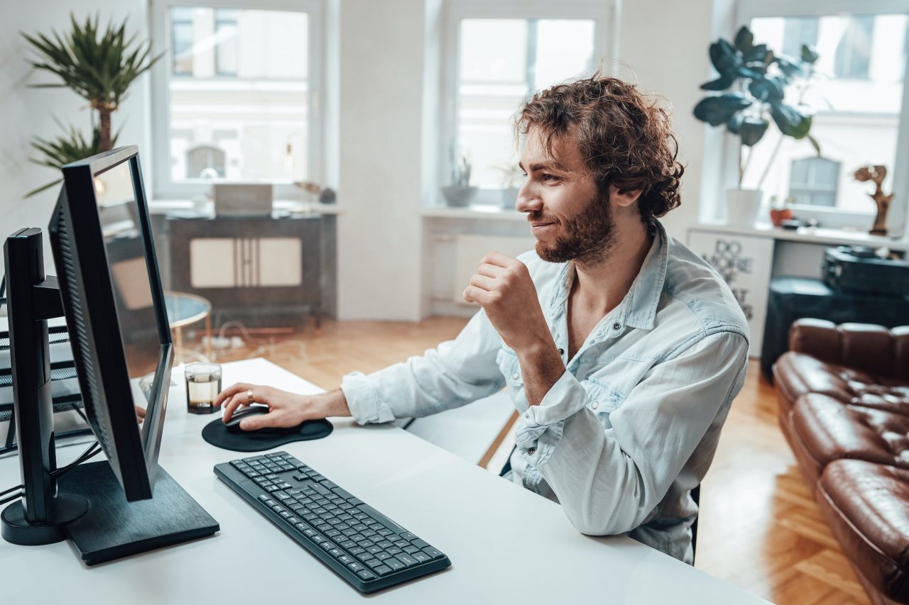 joyful guy with curly hairs and beard sits at table working on pc